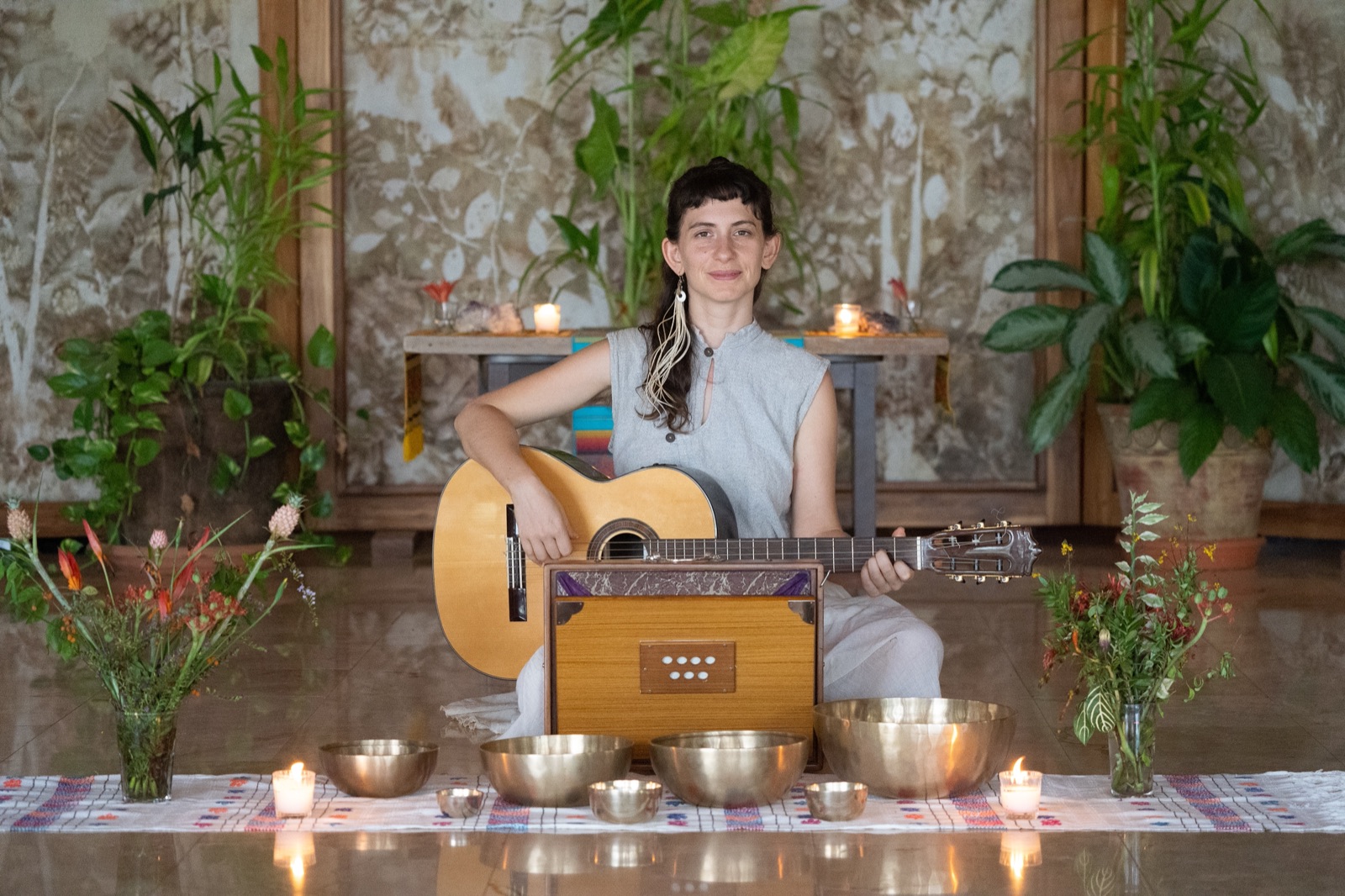 Yuli with a guitar behind a low altar of candles, singing bowls and plants